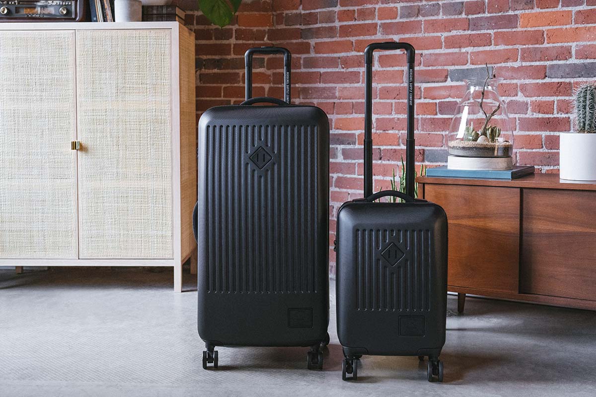 Un Trade Luggage Large nero & un Trade Luggage Carry-On uno accanto all’altro in un appartamento con mattoni a vista