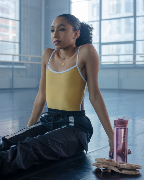 a woman sitting in a ballet studio with ballet shoes and a pink herschel water bottle next to her.