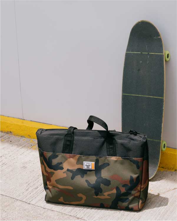a black/camo insulated alexander zip tote sits in front of a skateboarde leaning against a wall