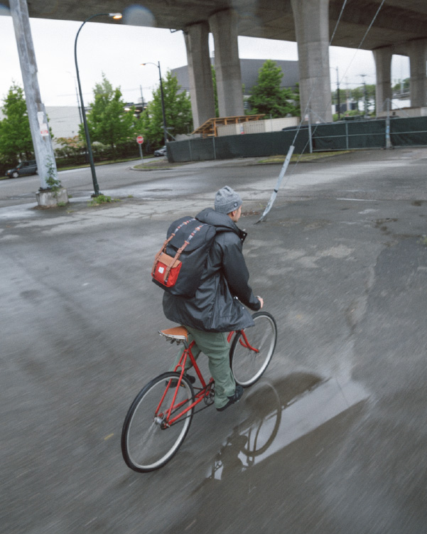 A person riding their bike under metro tracks while wearing their Herschel Little America Backpack.